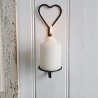 A white pillar candle in a black metal heart-shaped wall sconce, lit with a visible flame against a white paneled wall.