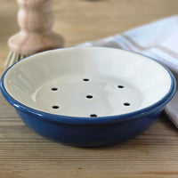 A small round enamel soap dish with a blue exterior and a white interior featuring five drainage holes, shown on a wooden tabletop with a blurred background.