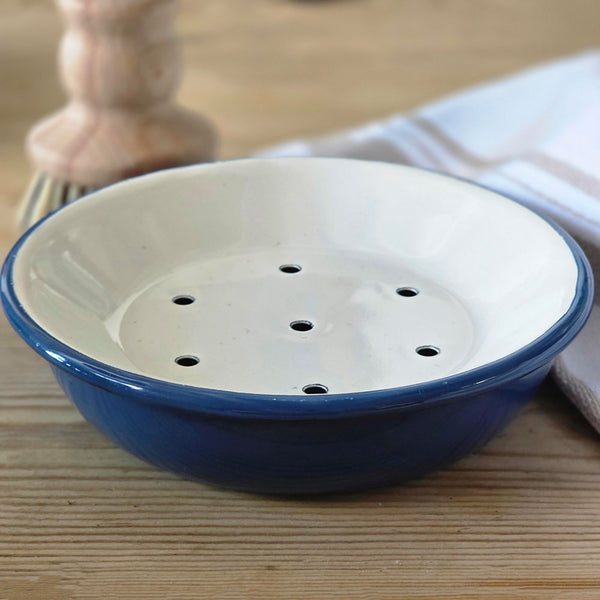 A small round enamel soap dish with a blue exterior and a white interior featuring five drainage holes, shown on a wooden tabletop with a blurred background.