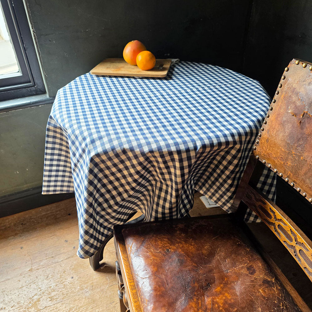 Round table with a blue and white checkered tablecloth, wooden bench, and oranges on a windowsill.