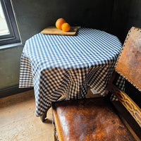 Round table with a blue and white checkered tablecloth, wooden bench, and oranges on a windowsill.