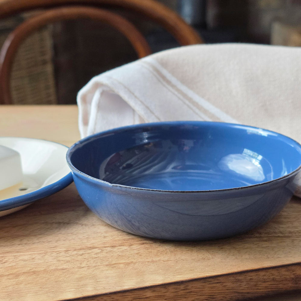 A blue enamel drainage tray with a glossy finish, shown on a wooden table next to a white dish with a blue rim and a folded cream linen cloth.