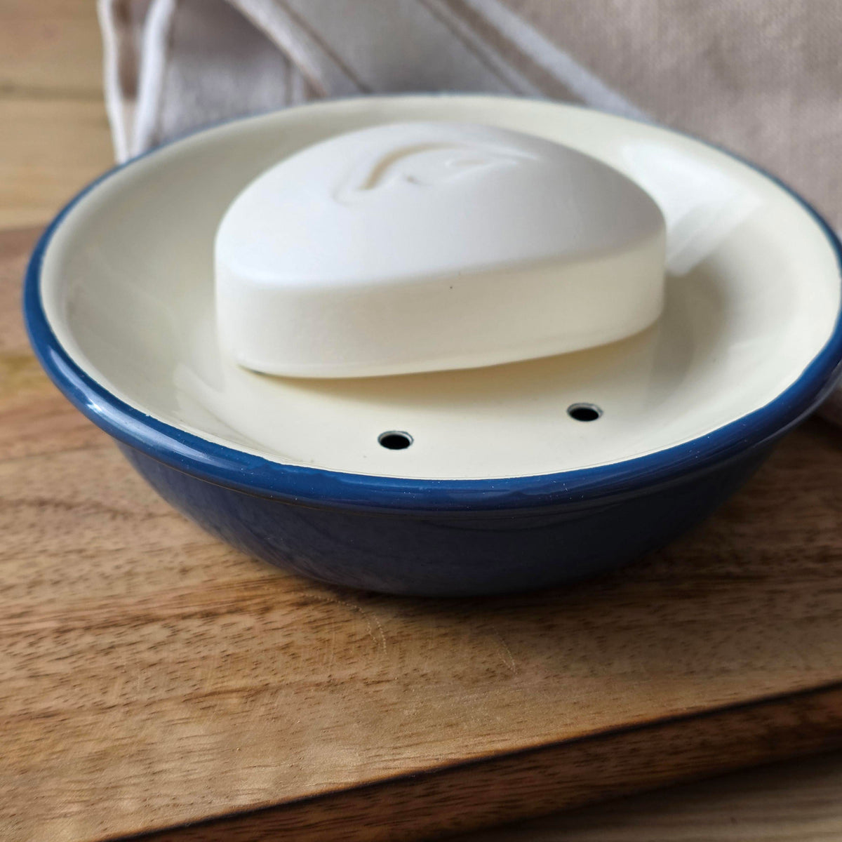 A ceramic soap dish with a blue rim and cream interior, holding a white bar of soap, set on a wooden surface with a soft neutral cloth in the background.