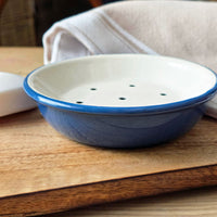 A small round enamel soap dish with a blue rim and base, and a white interior with several drainage holes, shown on a wooden surface with a folded cloth in the background.