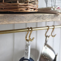 A close-up of brass kitchen hooks hanging from a brass rod on a rustic wooden shelf, with a wicker basket and kitchen items in the background.