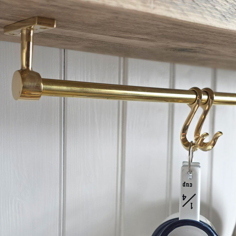 A close-up of a brass hanging rod mounted under a wooden shelf, with two decorative hooks holding a white cup against a white paneled wall.