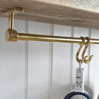 A close-up of a brass hanging rod mounted under a wooden shelf, with two decorative hooks holding a white cup against a white paneled wall.