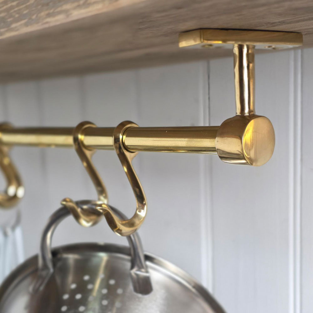 A close-up of a polished brass over-the-shelf hanging rod with hooks, shown mounted under a wooden shelf with a metal colander hanging from one hook.