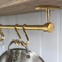 A close-up of a polished brass over-the-shelf hanging rod with hooks, shown mounted under a wooden shelf with a metal colander hanging from one hook.