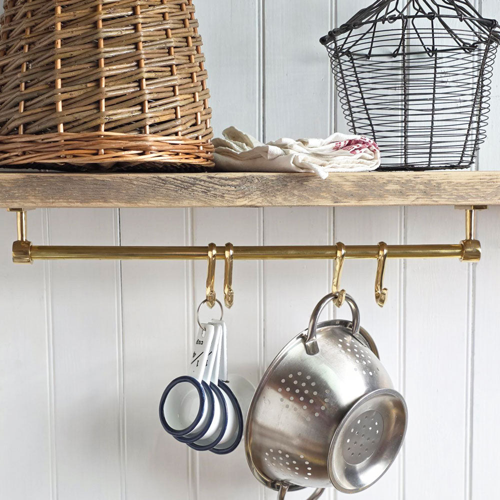 A brass kitchen wall rack with hanging pots and pans, shown installed on a wooden shelf in a kitchen setting with a wicker basket and white paneled wall.