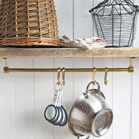 A brass kitchen wall rack with hanging pots and pans, shown installed on a wooden shelf in a kitchen setting with a wicker basket and white paneled wall.