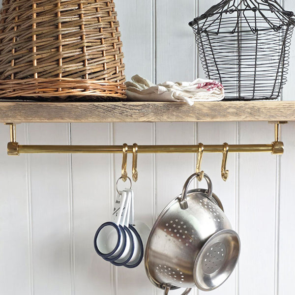 A brass kitchen wall rack with hanging pots and pans, shown installed on a wooden shelf in a kitchen setting with a wicker basket and white paneled wall.