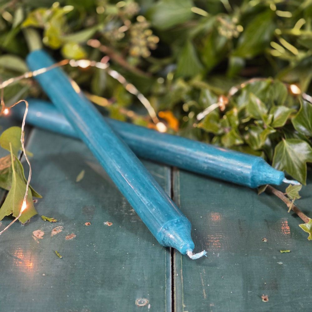 Two petrol blue candles on a wooden surface with green foliage in the background