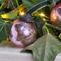 A close-up of a decorative glass Christmas ornament with a metallic pink tint and a dark metal cap, nestled among green ivy leaves and warm yellow string lights.