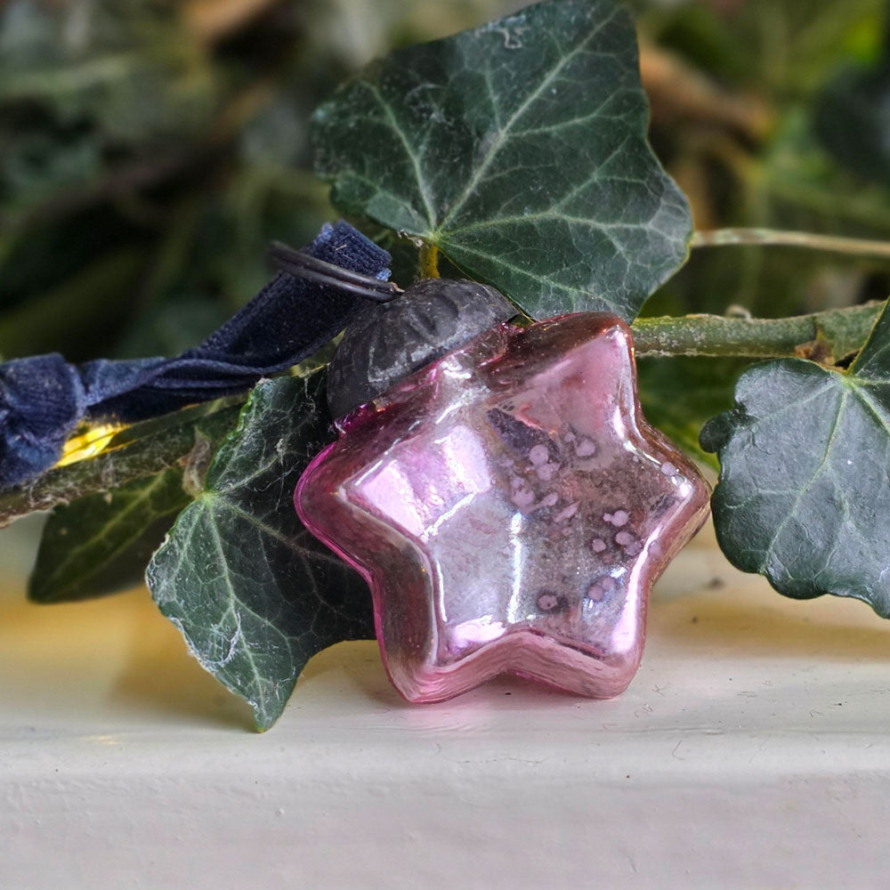 A star-shaped pink glass ornament with embedded glitter, resting on green ivy leaves with a dark blue ribbon attached.