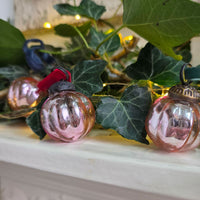 Three vintage-style pink glass Christmas ornaments with red, green, and bronze hanging tabs, displayed on a white mantelpiece among green ivy and warm fairy lights.