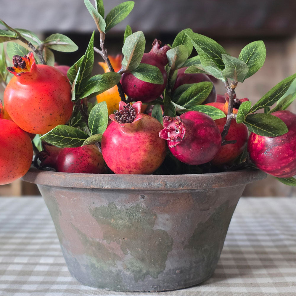 A decorative artificial pomegranate arrangement in a rustic distressed metal pot, featuring realistic red and pink pomegranates with green leaves and stems, set on a checkered tablecloth.