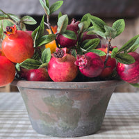 A decorative artificial pomegranate arrangement in a rustic distressed metal pot, featuring realistic red and pink pomegranates with green leaves and stems, set on a checkered tablecloth.