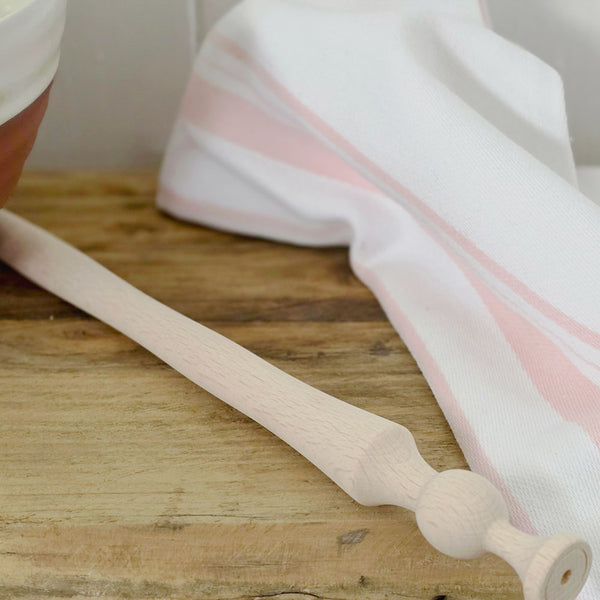 Wooden porridge spurtle on a wooden surface with a white and pink striped tea towel in the background.