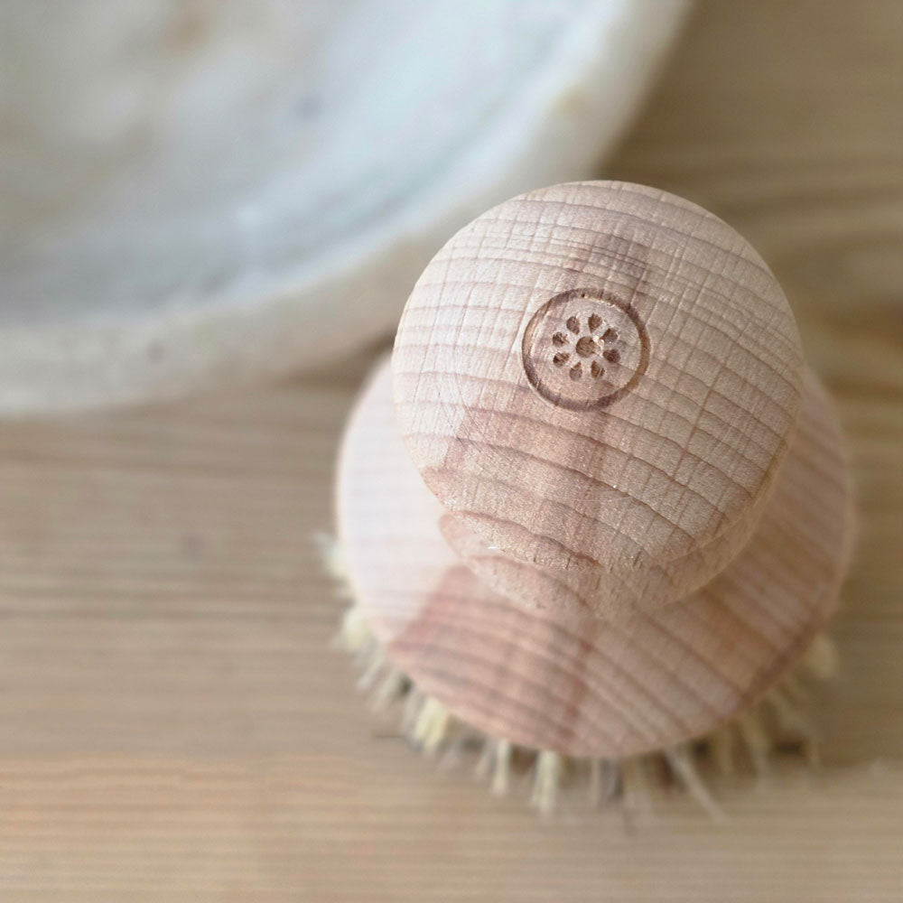 A close-up of a light natural wood scrubber brush with a carved handle and soft bristles, resting on a wooden surface.