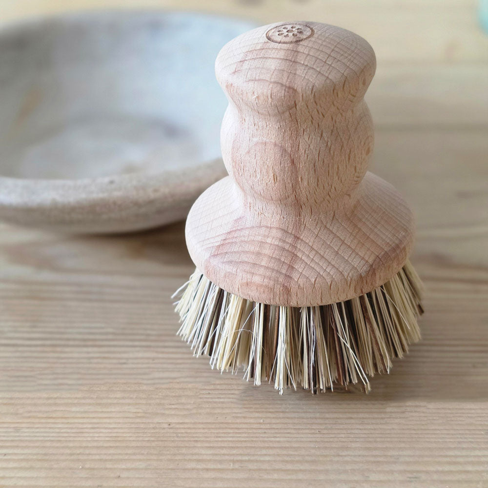 A close-up of a wooden dishwashing brush with a natural wood handle and natural bristles, standing on a light wood surface with a ceramic bowl blurred in the background.