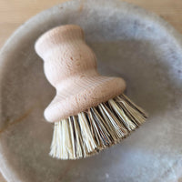 A close-up of a small hand brush with a light natural wood handle and beige bristles, resting inside a round gray stone bowl on a wooden surface.
