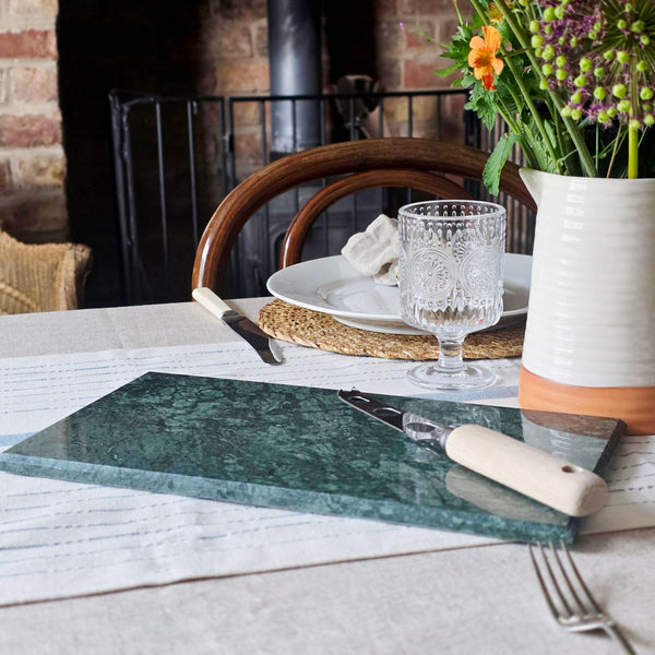 Dining table setting with a green marble cutting board, glass, and flowers in a vase.