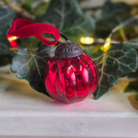 A small red glass Christmas ornament with a dark metal cap and a red ribbon loop, photographed among green ivy leaves and warm string lights.