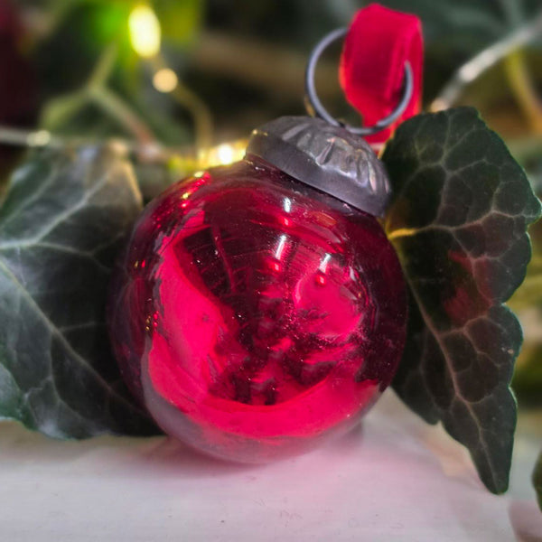 A red glass Christmas ornament bauble with a silver metal cap and red hanging loop, nestled among dark green ivy leaves.
