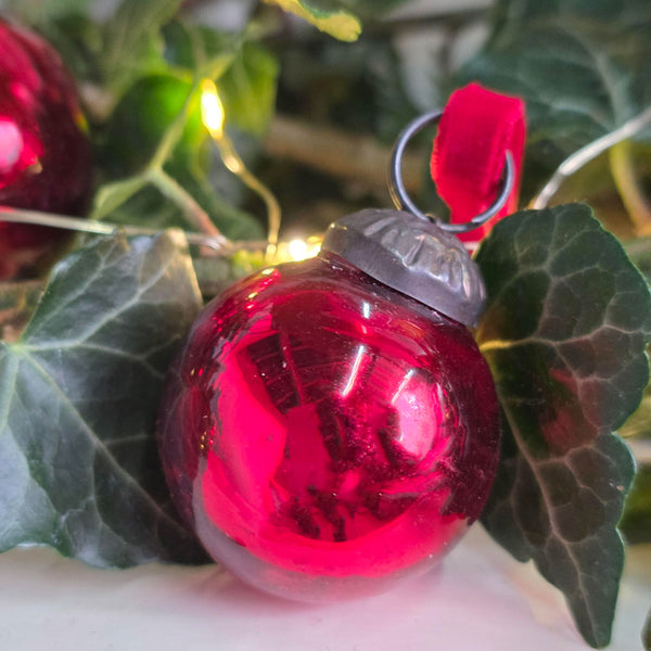 A glossy red Christmas bauble ornament with a silver metal cap and loop, nestled among green ivy leaves and warm string lights.