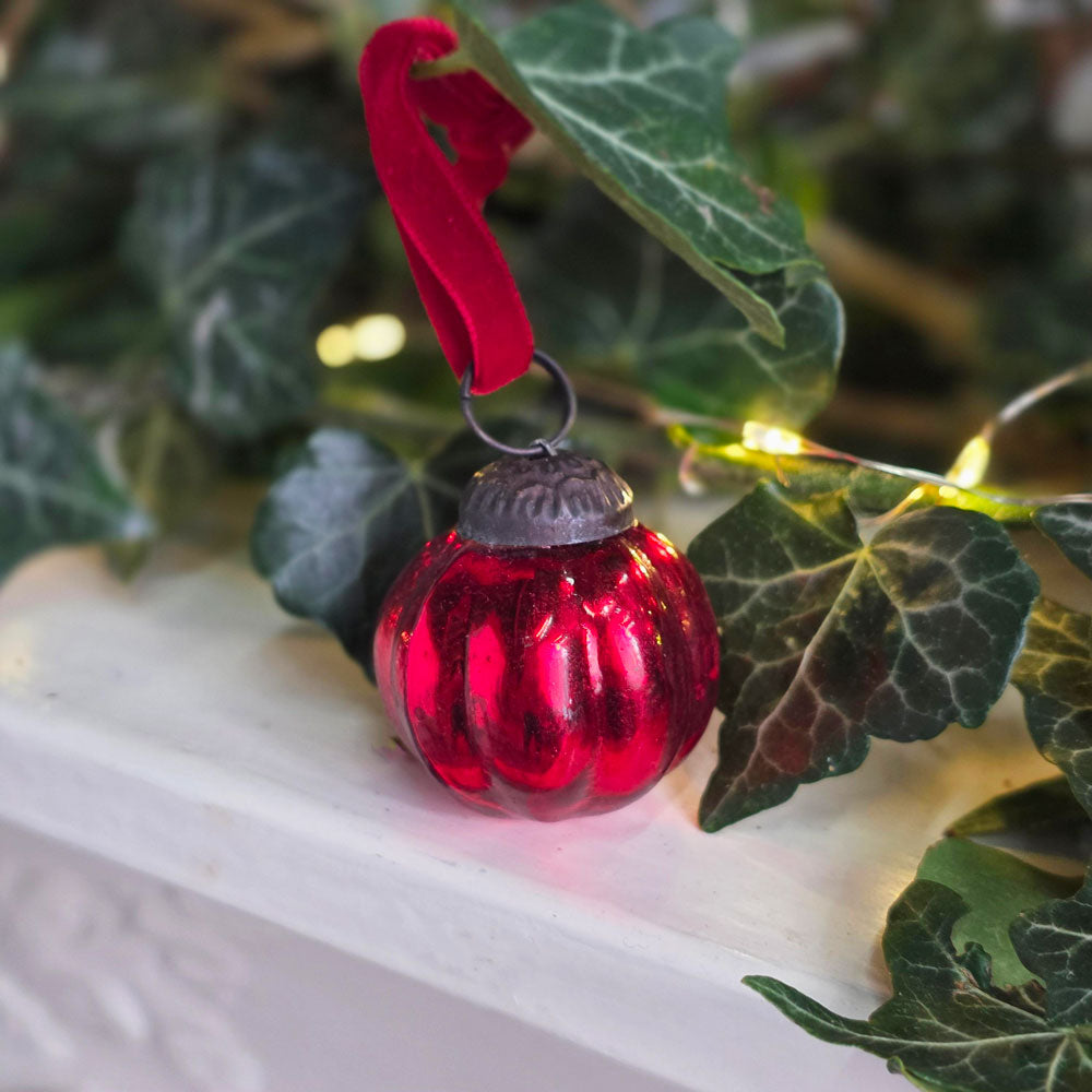 A small red glass Christmas ornament with a red velvet ribbon hanger, resting on a white mantel surrounded by green ivy leaves and warm string lights.