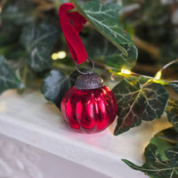 A small red glass Christmas ornament with a red velvet ribbon hanger, resting on a white mantel surrounded by green ivy leaves and warm string lights.