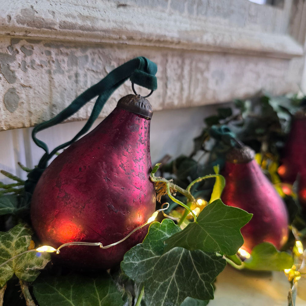 A close-up of a pear-shaped Christmas ornament in metallic burgundy with a dark green velvet hanging ribbon, nestled among green ivy and warm white string lights against a stone wall.