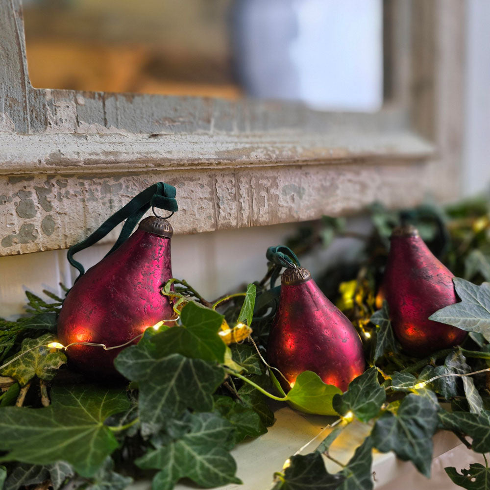 Three red pear-shaped Christmas ornaments with green ribbons and warm white string lights, nestled in green ivy garland near a rustic window frame.