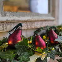 Three red pear-shaped Christmas ornaments with green ribbons and warm white string lights, nestled in green ivy garland near a rustic window frame.