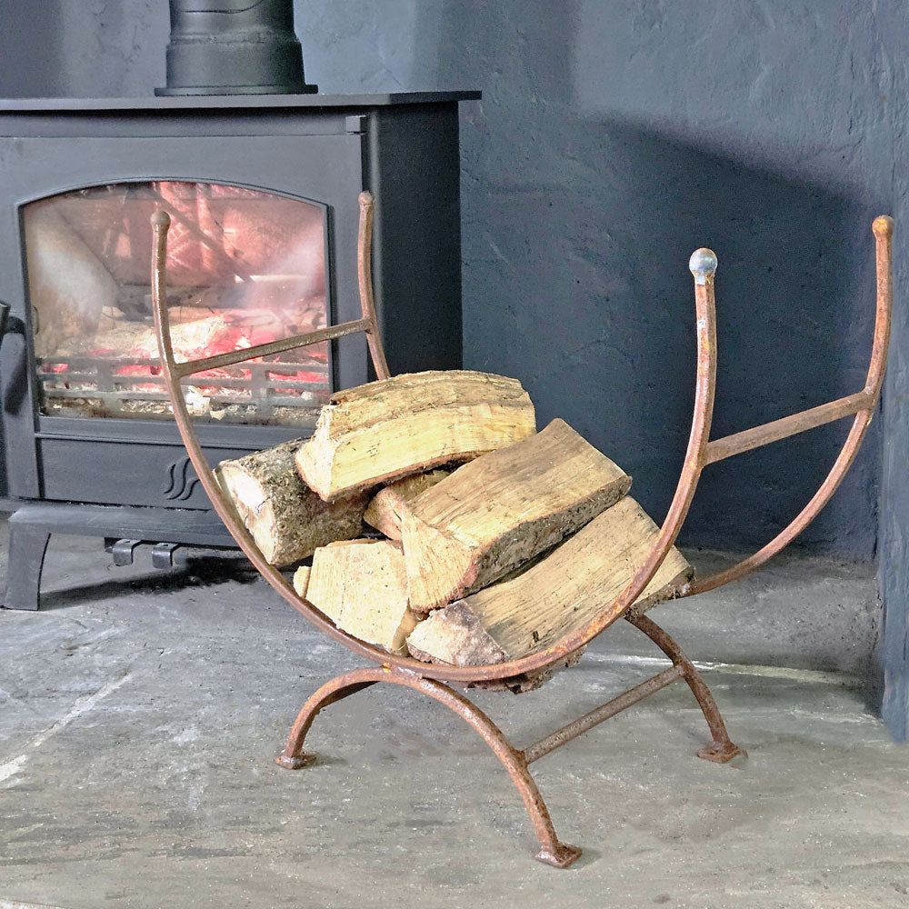 Traditional iron log holder standing on a fireside next to a lit wood burning stove.