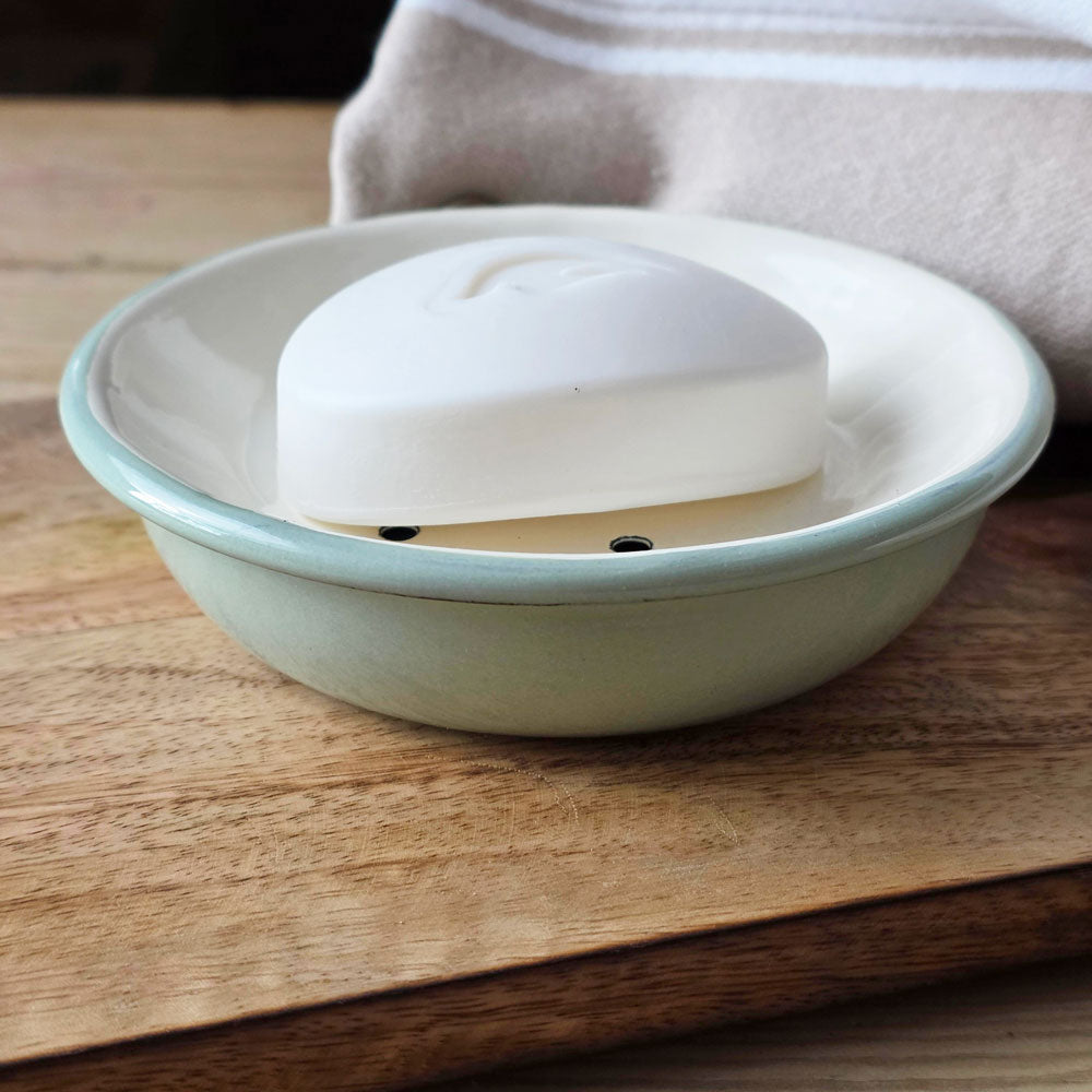 White bar of soap on a green and white circular enamel soap dish on a wooden board next to a white and beige towel.