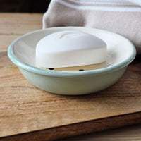 White bar of soap on a green and white circular enamel soap dish on a wooden board next to a white and beige towel.