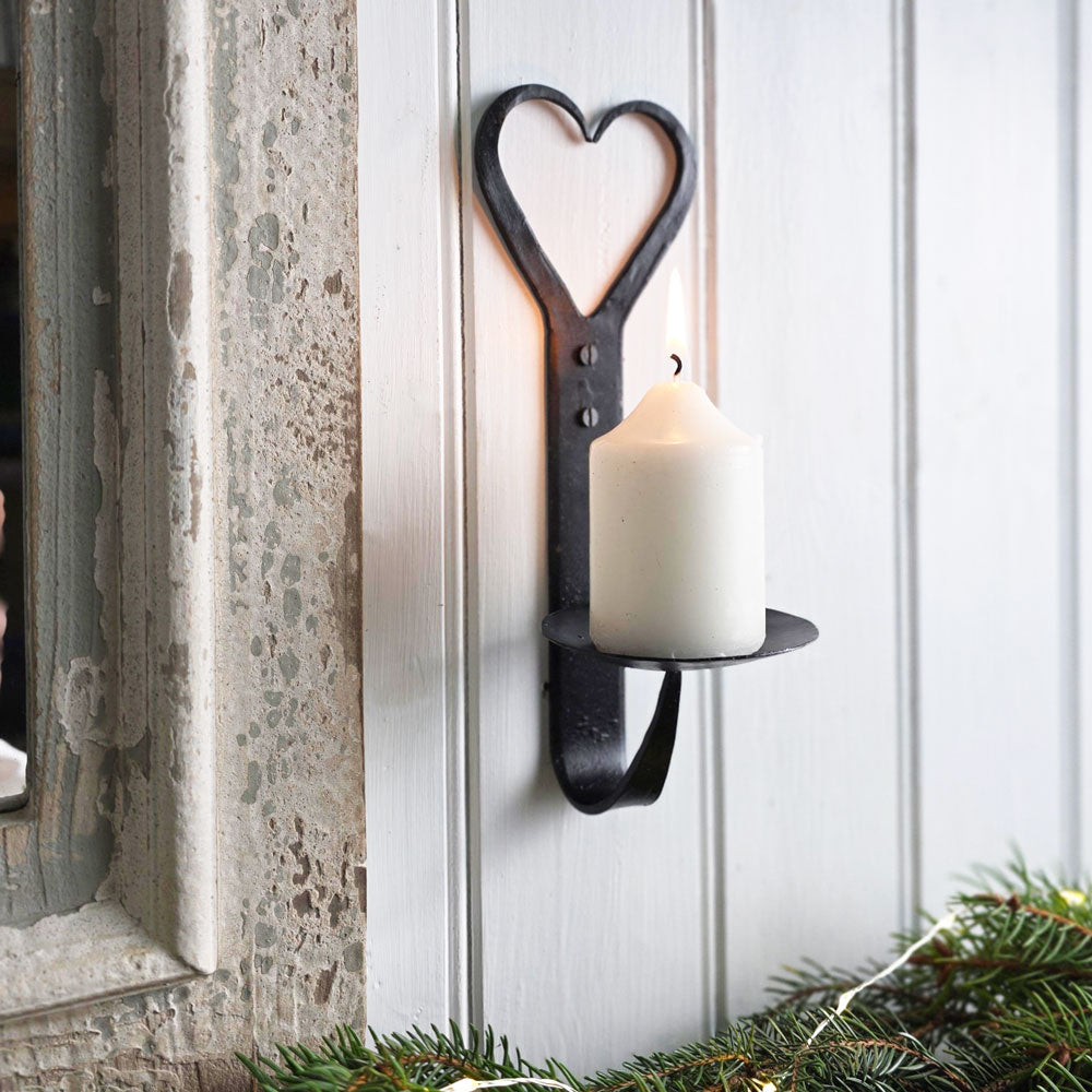 Forged iron wall mounted metal candle holder holding a small cream li candle next top a wooden frame with Christmas foliage.