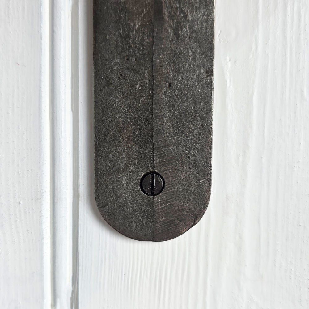 A close-up of a dark, weathered metal door hardware piece, likely a latch or bracket, secured with a central screw against a white painted wooden door background.