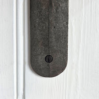 A close-up of a dark, weathered metal door hardware piece, likely a latch or bracket, secured with a central screw against a white painted wooden door background.