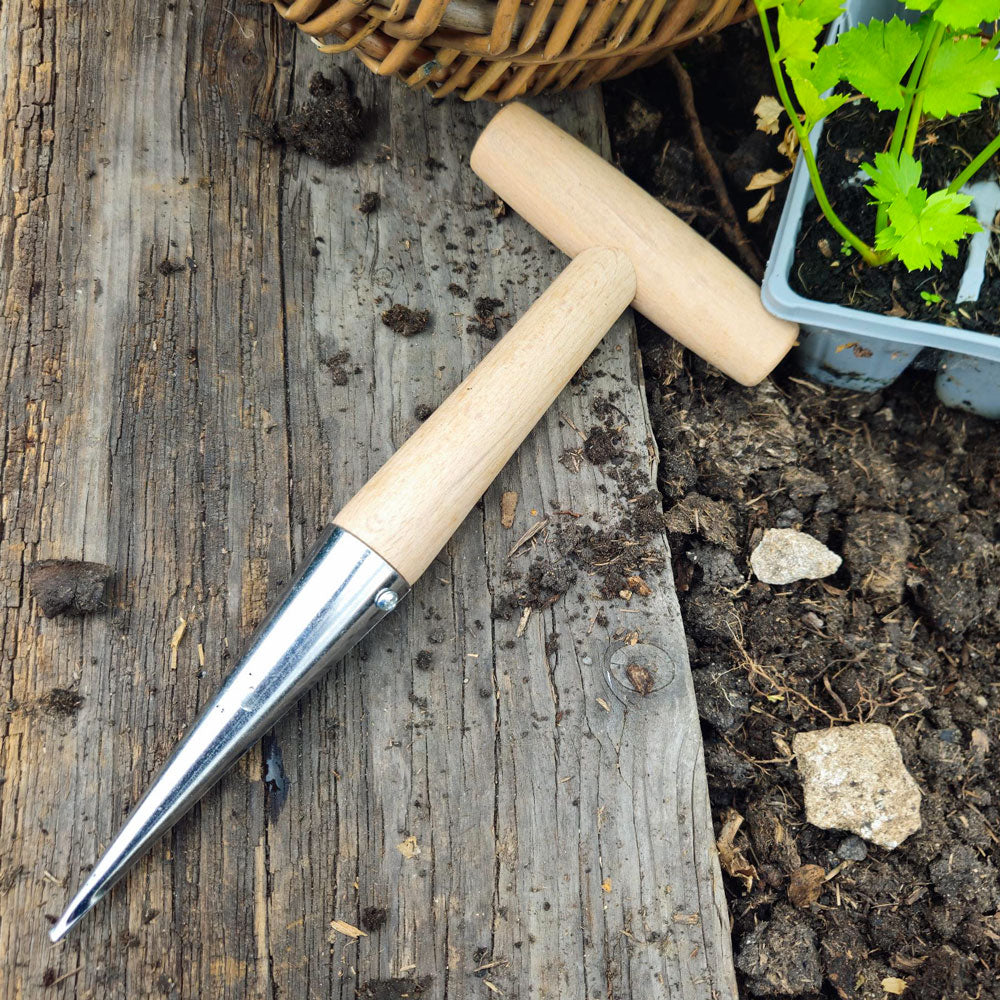 Garden dibber tool with wooden handle on a wooden surface with soil and plants.
