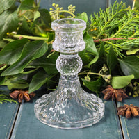 Clear glass candlestick on a wooden surface with greenery in the background