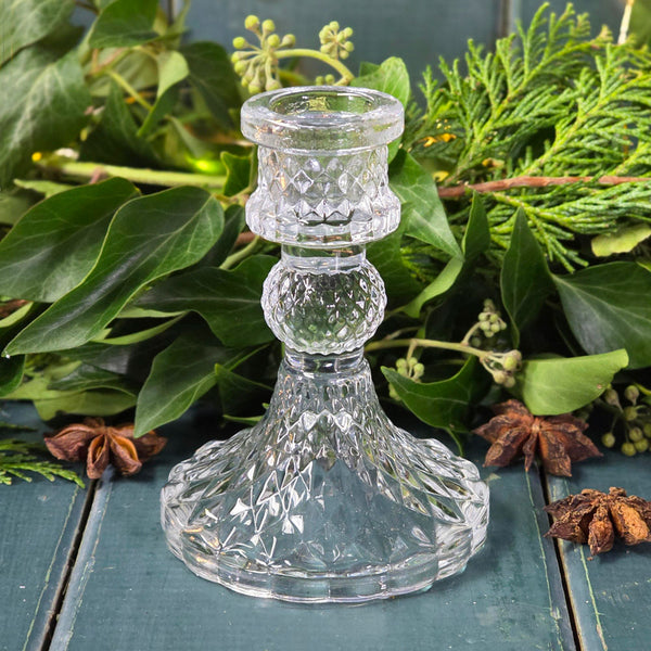 Clear glass candlestick on a wooden surface with greenery in the background