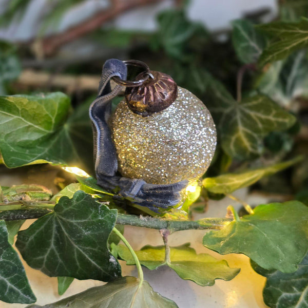 A glittery gold Christmas ornament with a dark gray ribbon and brown top loop, nestled among green ivy leaves with warm string lights.