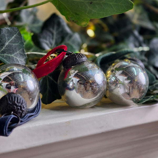 Three clear glass Christmas baubles with black caps, one with a red ribbon and one with a dark blue ribbon, resting on a white ledge surrounded by green ivy leaves.