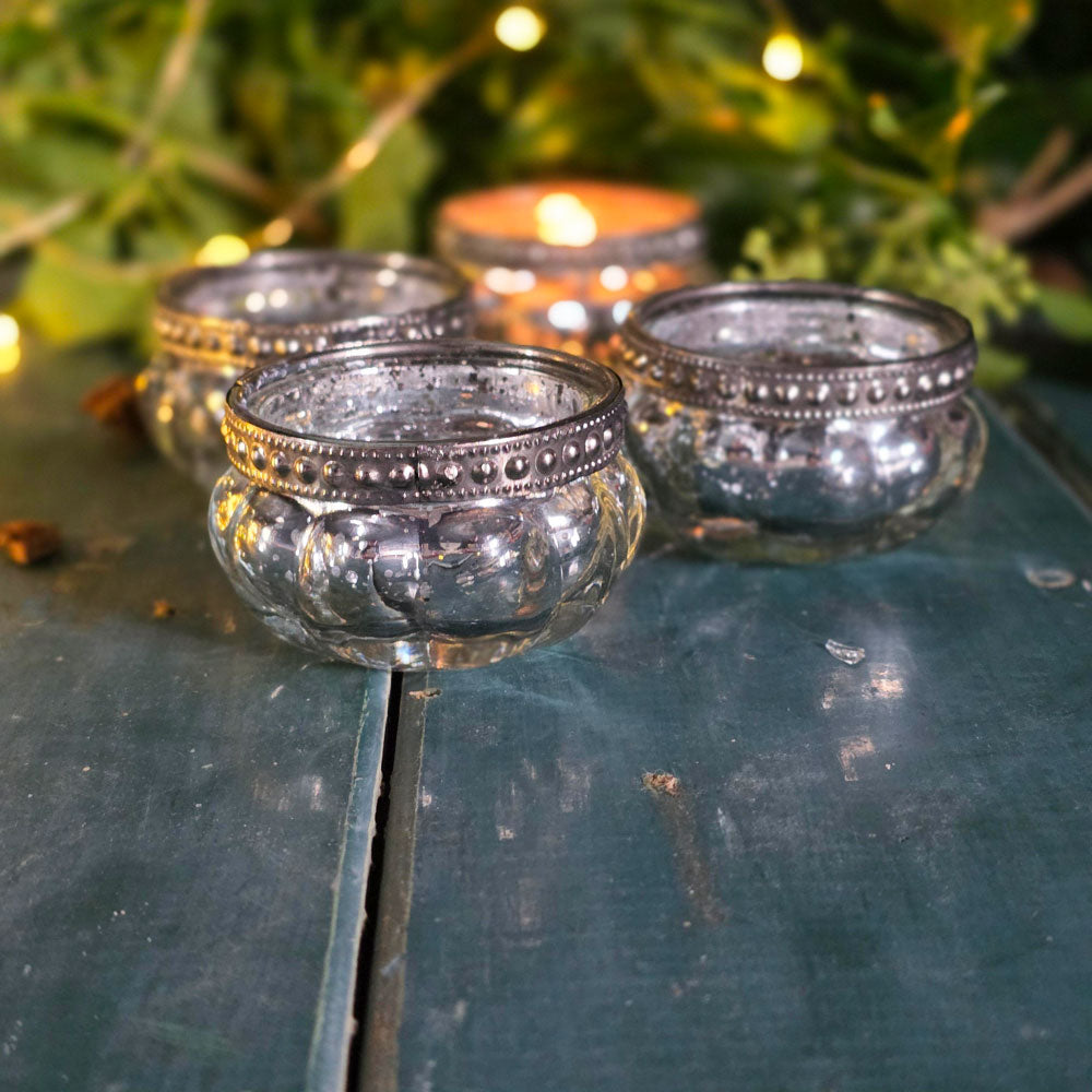 Decorative glass candle holders on a reflective surface with blurred greenery and lights in the background.