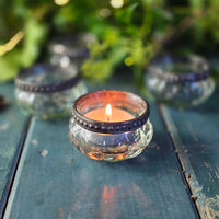 Candle in a decorative glass holder on a wooden surface with greenery in the background