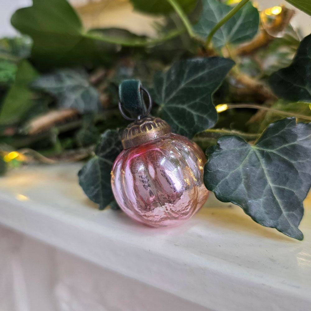 A small pink glass Christmas ornament with a ribbed texture and a dark green velvet loop, resting on a white mantel surrounded by green ivy leaves and warm string lights.