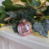 A small pink glass Christmas ornament with a ribbed texture and a dark green velvet loop, resting on a white mantel surrounded by green ivy leaves and warm string lights.
